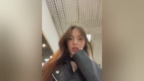 A video of a young woman with long brown hair, wearing a black leather jacket and black shirt, standing in a modern, beige-toned room with a grid ceiling.