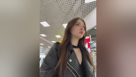 Video of a young woman with long brown hair, wearing a black choker and a grey jacket, standing in a modern shopping mall with a grid-patterned ceiling and fluorescent lighting.