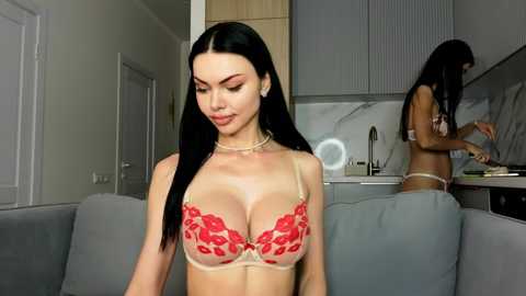 Video of a young woman with long black hair and fair skin in red lace bra, standing in modern kitchen with grey cabinets, marble backsplash, and white door.