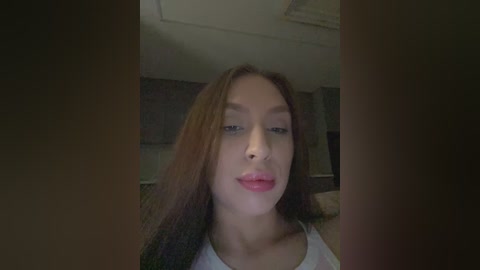 Video of a young Asian woman with long brown hair, light skin, and pink lipstick, wearing a white tank top, in a dimly lit room with beige walls and a ceiling fan.