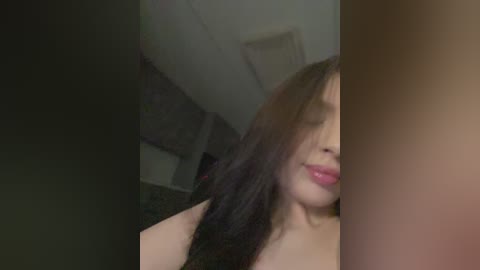 A video of a young woman with long, dark hair, partially covering her face, wearing red lipstick. She is indoors, with a dimly lit, carpeted room visible in the background.