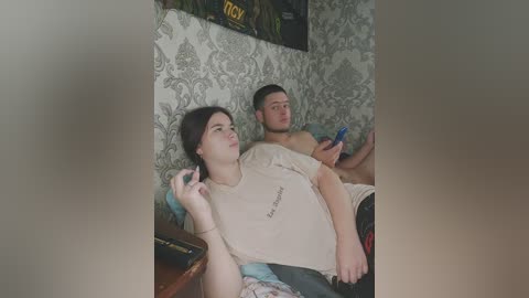 Video of a young couple watching TV in a retro-themed room with floral wallpaper and a poster. The man is shirtless, holding a remote, and the woman, wearing a T-shirt, is on the couch.
