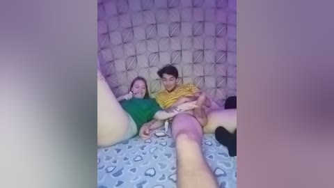 Video of a young couple in a dimly lit, retro-themed room with a tiled wall. The man is wearing a yellow shirt, and the woman is in a green top, both sitting on a patterned bedspread.