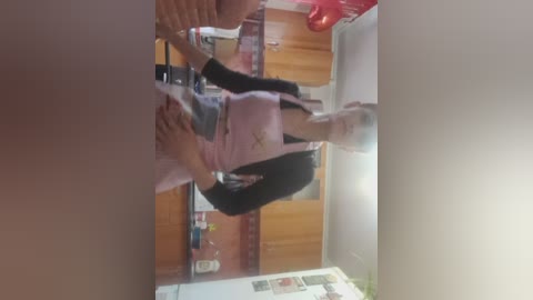 Video of a woman in a pink apron and black top, holding a child in a white shirt, standing in a kitchen with wooden cabinets and a fridge in the background.