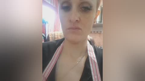 Video of a pale-skinned woman with short blonde hair, wearing a black top with red and white stripes, looking down with a neutral expression. Background includes a wooden kitchen counter and hanging utensils.