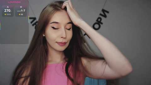 A video of a light-skinned woman with long brown hair, wearing a pink sleeveless top, smiling and touching her hair, against a gray wall with \"OPRAH\" and \"A\" text.