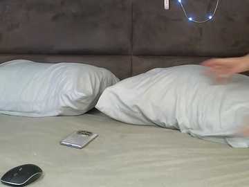 Video of a person in a bed with a dark grey headboard, holding a smartphone. Light grey pillows and duvet, a white mouse and charging cable on the bed, and a blue glowing light on the wall.