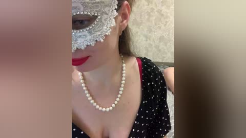 Video of a woman with fair skin, wearing a black polka-dotted dress, a pearl necklace, and a silver masquerade mask. The background features beige damask wallpaper.