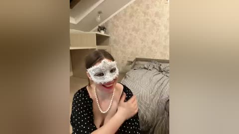 Video of a woman in a black polka-dot dress, wearing a white lace masquerade mask and pearl necklace, sitting on a bed with gray bedding. The room has beige wallpaper and a wooden shelf in the background.
