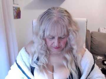 A video of a middle-aged woman with long, curly, platinum blonde hair, wearing a fluffy white bathrobe, seated indoors with a serene expression, and a beige sofa in the background.