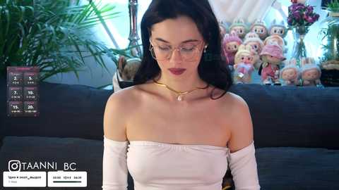 Video of a fair-skinned woman with long black hair, wearing glasses and a white off-shoulder top, seated on a dark couch, surrounded by plush toys.