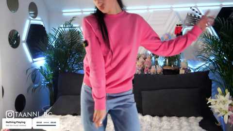 Video of a woman in a pink sweater and blue jeans, dancing in a modern, brightly lit room with plants and black furniture.