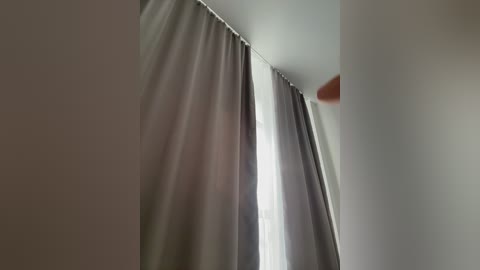 A video of a room featuring a window partially covered by gray and white curtains. The background is a plain, light-colored wall. The image is taken from a low angle, emphasizing the curtains' texture and the soft light filtering through.