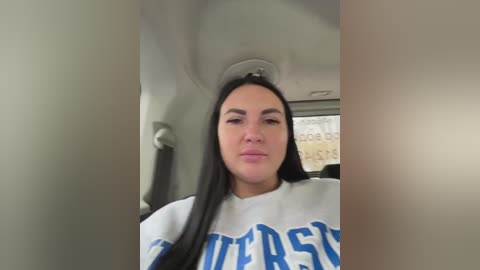 A video of a woman with long black hair, wearing a white \"Wolverines\" sweatshirt, seated in a car with beige walls and a large window displaying a cityscape.