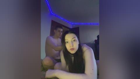 Video of an Asian woman with long black hair and a slender build, topless, sitting on a couch, with a shirtless man behind her, both under blue LED strip lighting.
