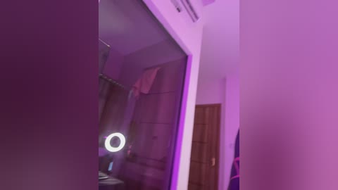 Video of a dimly lit bathroom with a purple hue, featuring a round mirror with a bright circular light, a shower, and a wooden door in the background.