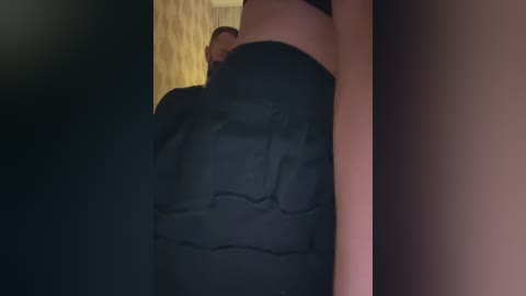 A video of a person, possibly male, lying on a bed with a black blanket partially covering their lower body, head obscured by the blanket, wooden floor visible in the background.