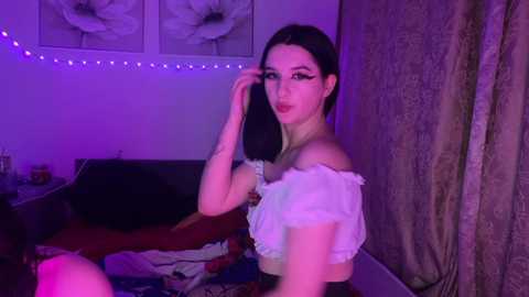 Video of a young woman with light skin, dark hair, and black eyeliner, wearing a white off-shoulder blouse and black bra. She is in a dimly lit bedroom with purple lighting, floral wallpaper, and a bed with red sheets.