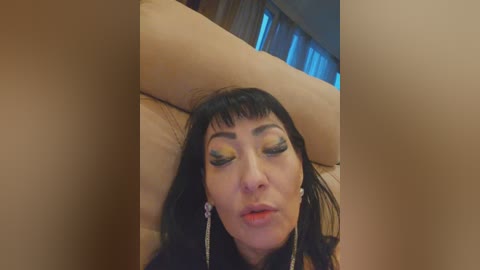 Video of a woman with long black hair, closed eyes, wearing dramatic green eyeshadow and dangling earrings, lying on a beige sofa with a blurred background.