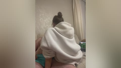 Video of a woman in a white bathrobe, sitting on a man's lap, facing away from the camera, in a dimly lit bedroom with cream-colored walls.