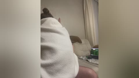 A video showing a woman with light skin, dark hair in a bun, wrapped in a white towel, sitting on a beige sofa, watching TV.