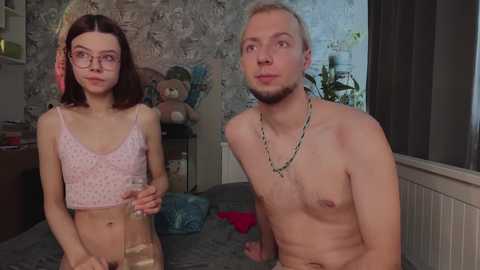 Video of a young, slender, pale-skinned woman with glasses and a pink polka-dot crop top, standing next to a shirtless, lean, fair-skinned man with a beard, both indoors.