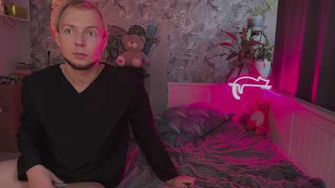 Video of a young man with light skin and short blonde hair, wearing a black V-neck shirt, sitting on a bed in a dimly lit room with neon lights.