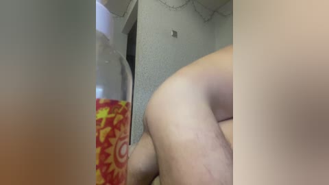 A video of a man's bare buttocks, with a red and yellow patterned bottle partially visible in the foreground. The background features a beige wall and a doorway.
