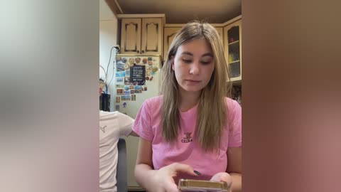 Video of a young Caucasian girl with straight blonde hair, wearing a pink t-shirt, holding a bottle in a kitchen with wooden cabinets, refrigerator, and magnets.
