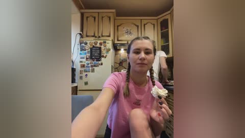 Video of a young Caucasian woman with light skin, brown hair in braids, wearing a pink t-shirt and holding a flower, sitting in a cozy kitchen with wooden cabinets and a refrigerator covered in magnets.