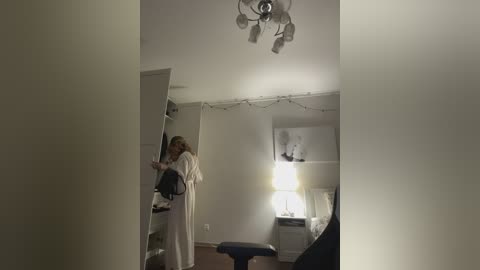 A dimly lit video of a woman in a white robe, holding a black purse, standing in a sparse, white-walled room with a chandelier, string lights, and a small desk with a lamp.