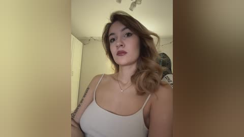 A video of a young woman with fair skin and wavy brown hair, wearing a white spaghetti-strap top, standing in a dimly lit, beige-walled room.