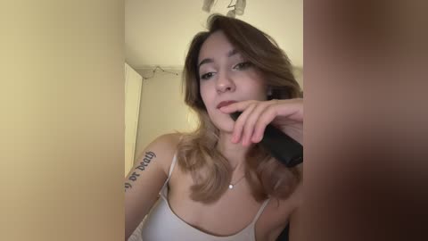 A video of a young woman with long, wavy brown hair, wearing a white spaghetti-strap tank top, sitting in a dimly-lit room, holding a black phone to her lips.