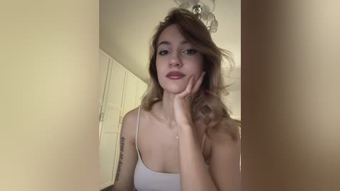 A video of a young, light-skinned woman with wavy brown hair, wearing a white spaghetti-strap tank top, sitting in a beige room with a white closet and ceiling fan.