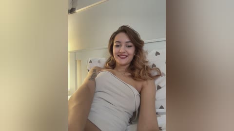 Video of a young woman with light skin, shoulder-length wavy brown hair, and red lipstick, wearing a beige strapless top, smiling in a room with a white bedspread and patterned pillows.