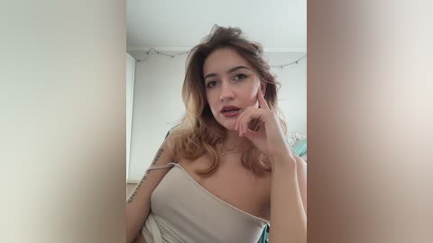 A young woman with fair skin and long, wavy blonde hair, wearing a strapless white top, poses thoughtfully indoors, with white walls and string lights in the background.