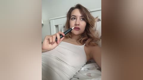 A video of a young woman with light skin and wavy brown hair, wearing a white tank top, applying lip gloss in a bedroom with light-colored bedding.
