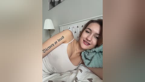 A video of a young woman with light skin and brown hair, wearing a white tank top, lying on a bed. She has a tattoo reading \"Blessing in Disguise\" on her arm. The room has a white wall and a lamp.