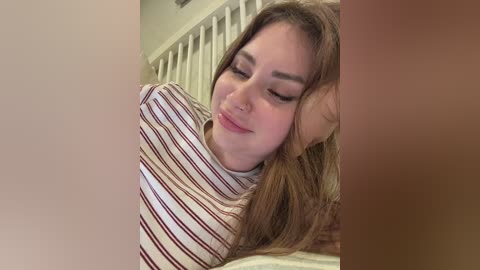 Video of a young woman with light skin, long brown hair, wearing a striped t-shirt, resting her head on her hand, smiling softly, in a white crib.