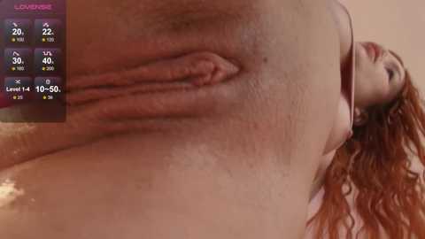 Video of a close-up, partially blurred, reddish-brown female vulva with visible labia and a hint of pubic hair. Background shows a partially visible, fair-skinned woman with wavy, auburn hair, and a pink top.