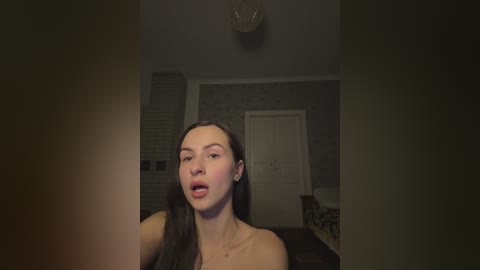 Video of a pale, dark-haired woman with a neutral expression, wearing a strapless black top, in a dimly lit room with beige walls and a door.