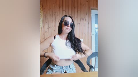 Video of a young woman with long black hair, wearing dark sunglasses, a white crop top, and ripped denim shorts, standing in a rustic wooden room.