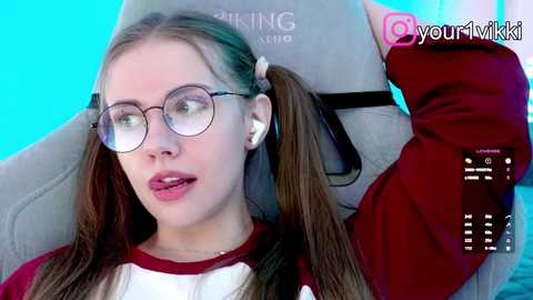 A video of a young woman with pigtails, wearing glasses, a red hoodie, and white shirt, lying on a grey gaming chair with a blue background.