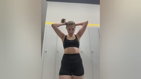 Video of a young woman with light skin and brown hair in a high ponytail, wearing a black sports bra and high-waisted shorts, standing in a locker room with gray lockers and a yellow stripe on the wall.