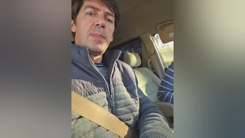 A video of a middle-aged man with short, dark hair, wearing a blue puffer jacket, driving a car with beige seats and blue patterned pillows in the background.
