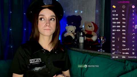 Video of a young Caucasian woman in a black police uniform, hat, and badge, standing in a dimly lit room with plush toys and a green curtain.