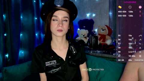 A video of a young, fair-skinned woman with short brown hair, wearing a black police hat and uniform, seated in a dimly lit room with blue fairy lights, teddy bears, and a 'PROTECTED' sign.