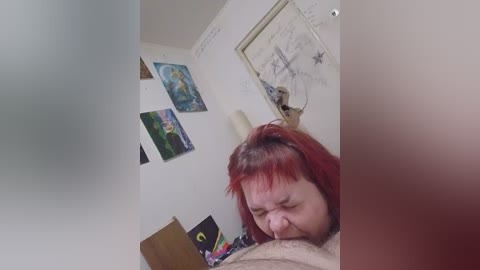 A video of a red-haired person with a medium build, wearing a beige shirt, kneeling on a bed, performing oral sex on a person's penis. The room has white walls adorned with colorful posters and a small desk.