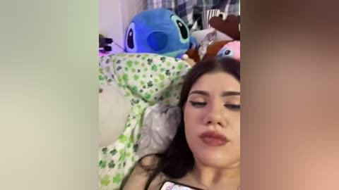 Video of a young woman with long dark hair, fair skin, and closed eyes lying on a bed with plush toys, including a blue Stitch and a green plush, in a cozy, cluttered room.