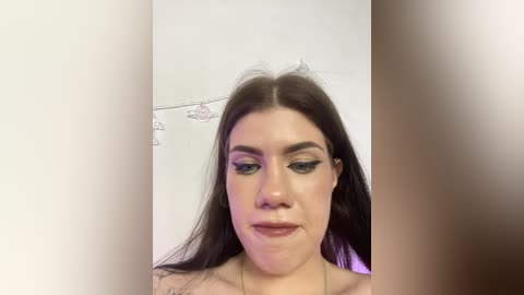 Video of a young woman with fair skin, dark hair, and green eyes, wearing dramatic eye makeup, looking into the camera. The background is a plain white wall.